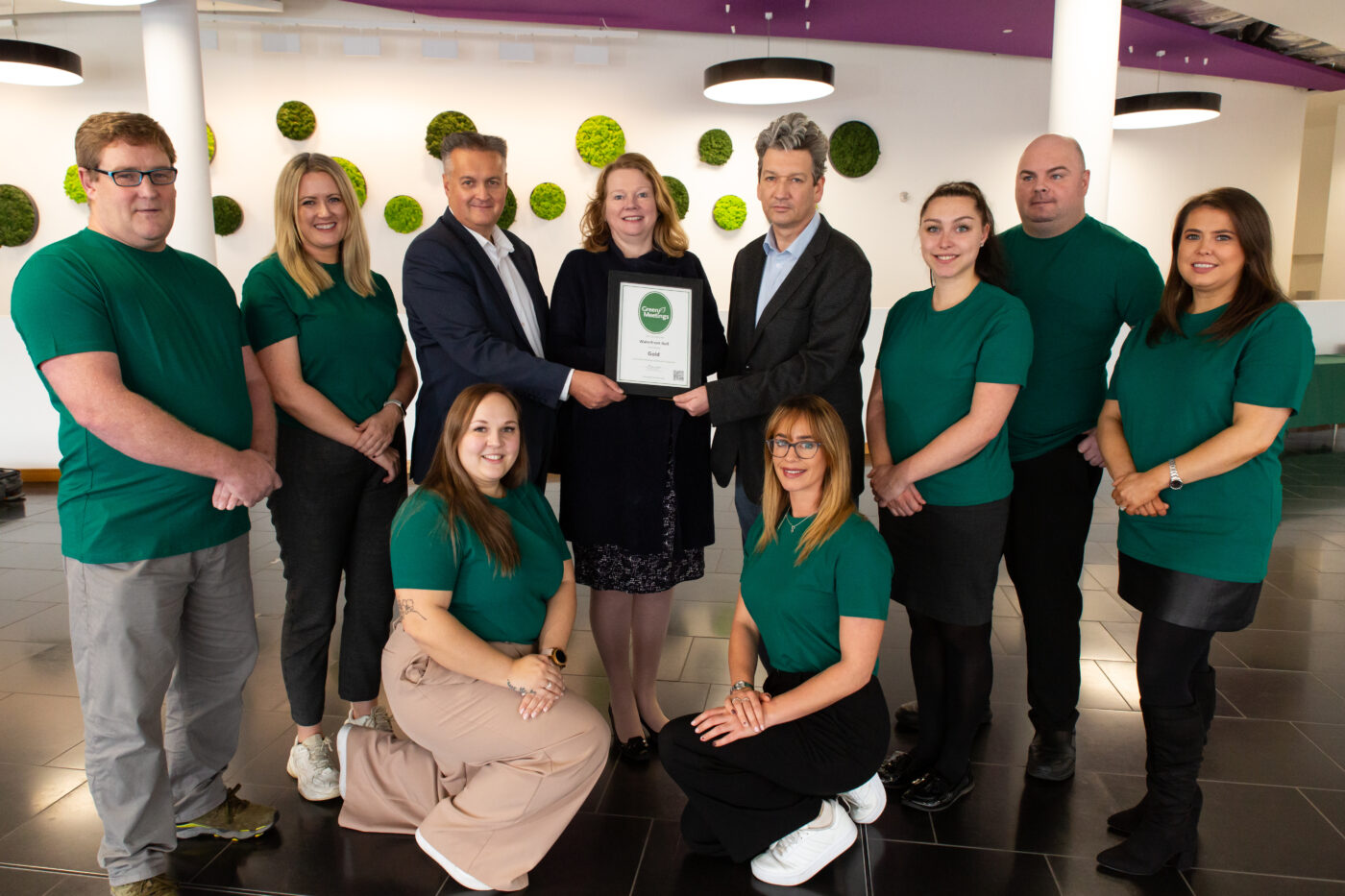 A group of ten people, some in green shirts, smile as two men and a woman in business attire hold an award certificate at the center. They are indoors with a green and white wall display behind them.